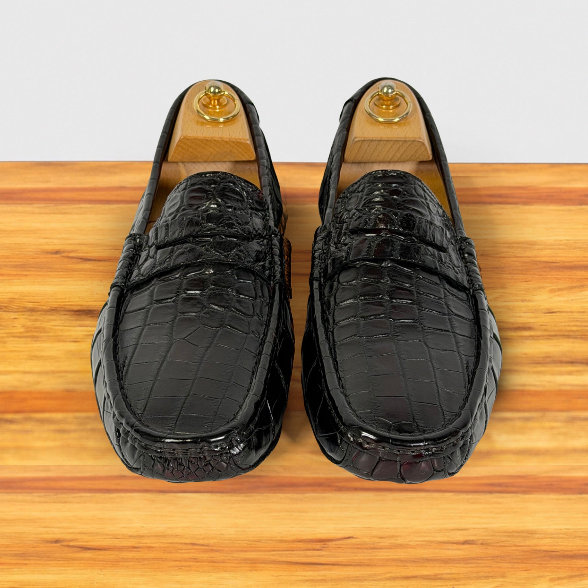A pair of burgundy crocodile-embossed leather loafers displayed on wooden shoe trees over a light wood surface against a white background.