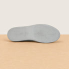 The outsole of a slip-on shoe, showing textured tread patterns, resting on a light wood surface against a beige background.