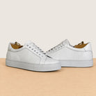 A pair of white leather low-top sneakers with perforated detailing, shown on a light wood surface against a warm beige background.