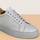 A white leather low-top sneaker with perforated side panels, matching white laces, and a thick textured sole, shown resting on a light wood surface against a cream background.