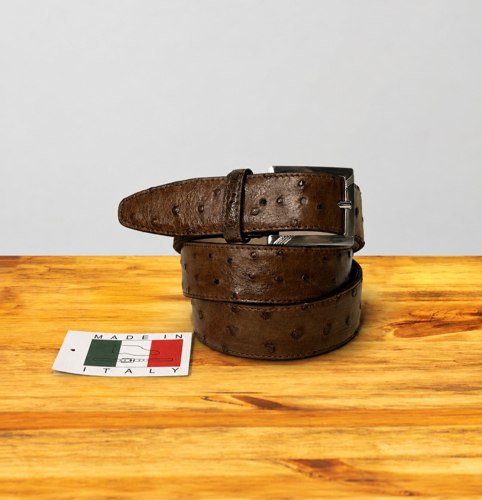 A cerris ostrich leather belt with a silver buckle, coiled on a wooden surface next to a small 'Made in Italy' tag, against a light gray background.