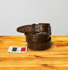 A cerris ostrich leather belt with a silver buckle, coiled on a wooden surface next to a small 'Made in Italy' tag, against a light gray background.