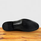 The smooth black leather outsole of a Dusty Brown dress shoe, shown from a bottom-up view resting on a wooden table.