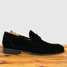 A single black suede penny loafer shown in profile on a wooden surface against a light gray background.