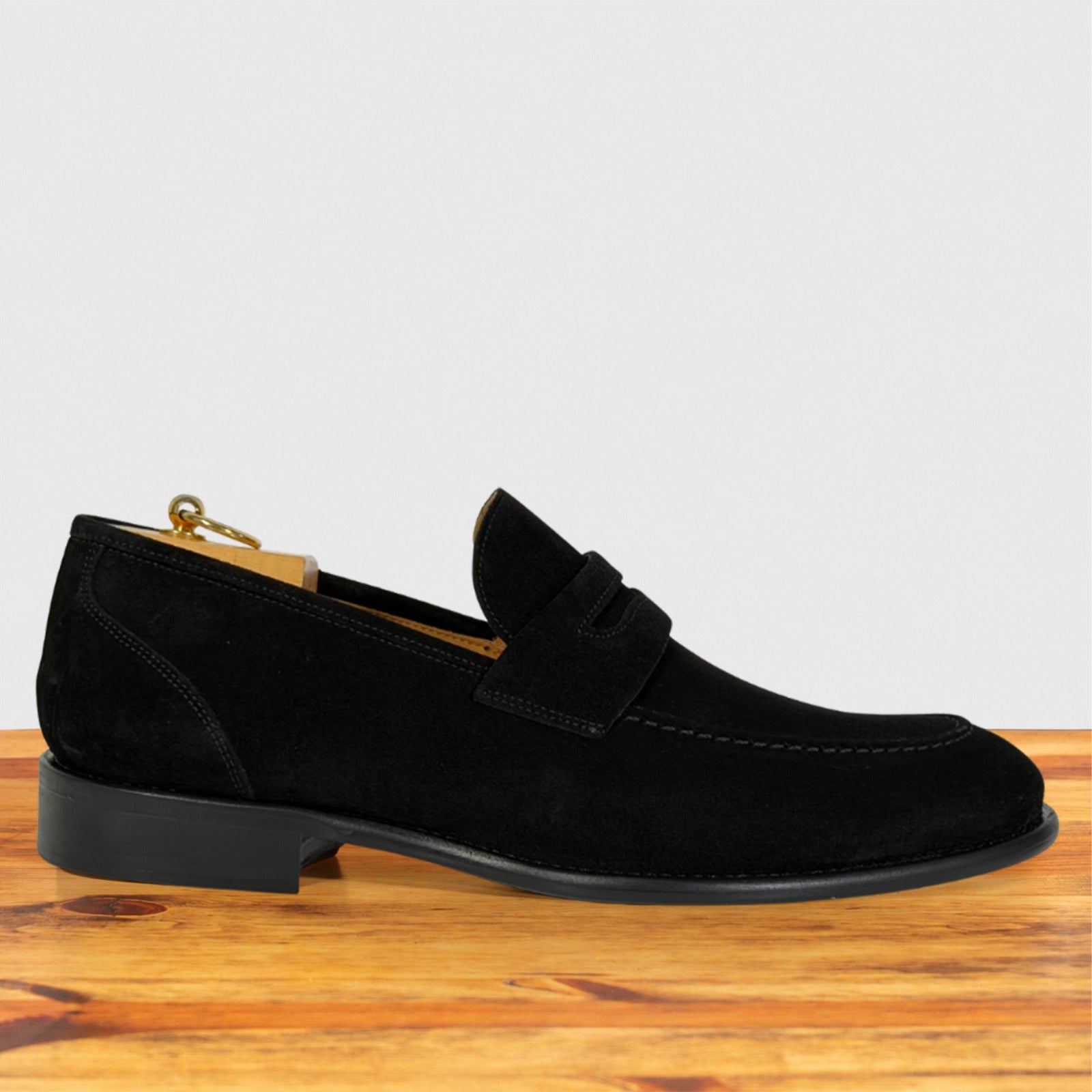 A single black suede penny loafer shown in profile on a wooden surface against a light gray background.