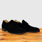 A pair of black suede penny loafers with low block heels, displayed on a wooden surface against a light gray background.