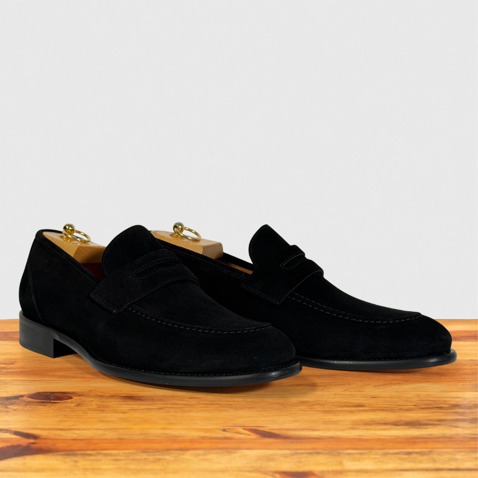 A pair of black suede penny loafers with low block heels, displayed on a wooden surface against a light gray background.