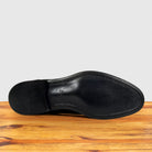 The black leather underside of a dress shoe sole, showing the smooth vamp and heel counter, resting on a wooden surface against a light gray background.