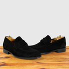 A pair of black suede penny loafers with low block heels, displayed on a wooden surface against a light gray background.
