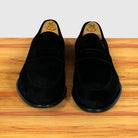 A pair of black suede penny loafers displayed, set on a wooden surface against a light gray background.