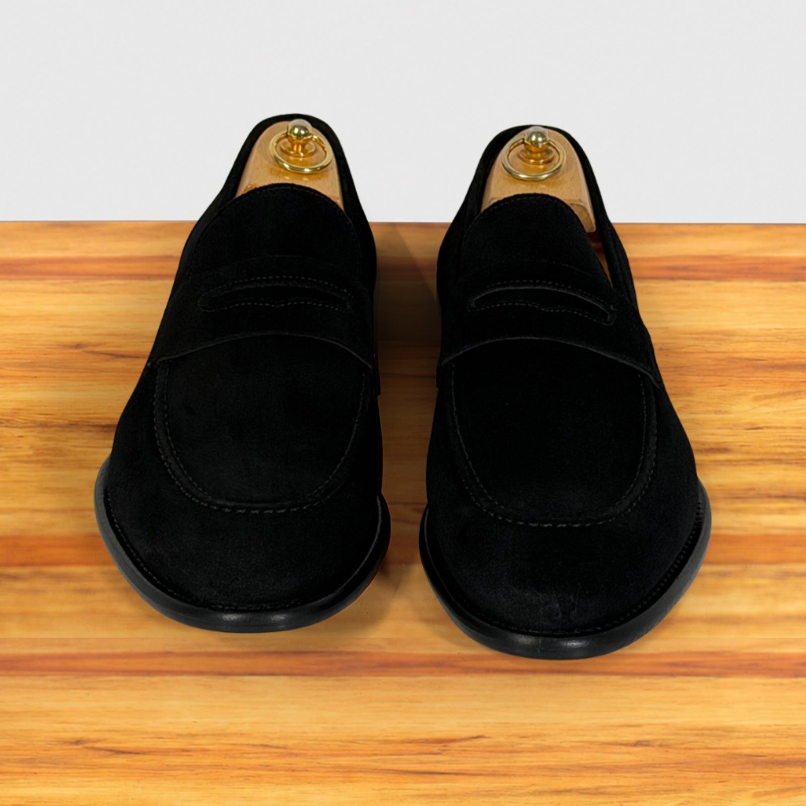 A pair of black suede penny loafers displayed, set on a wooden surface against a light gray background.