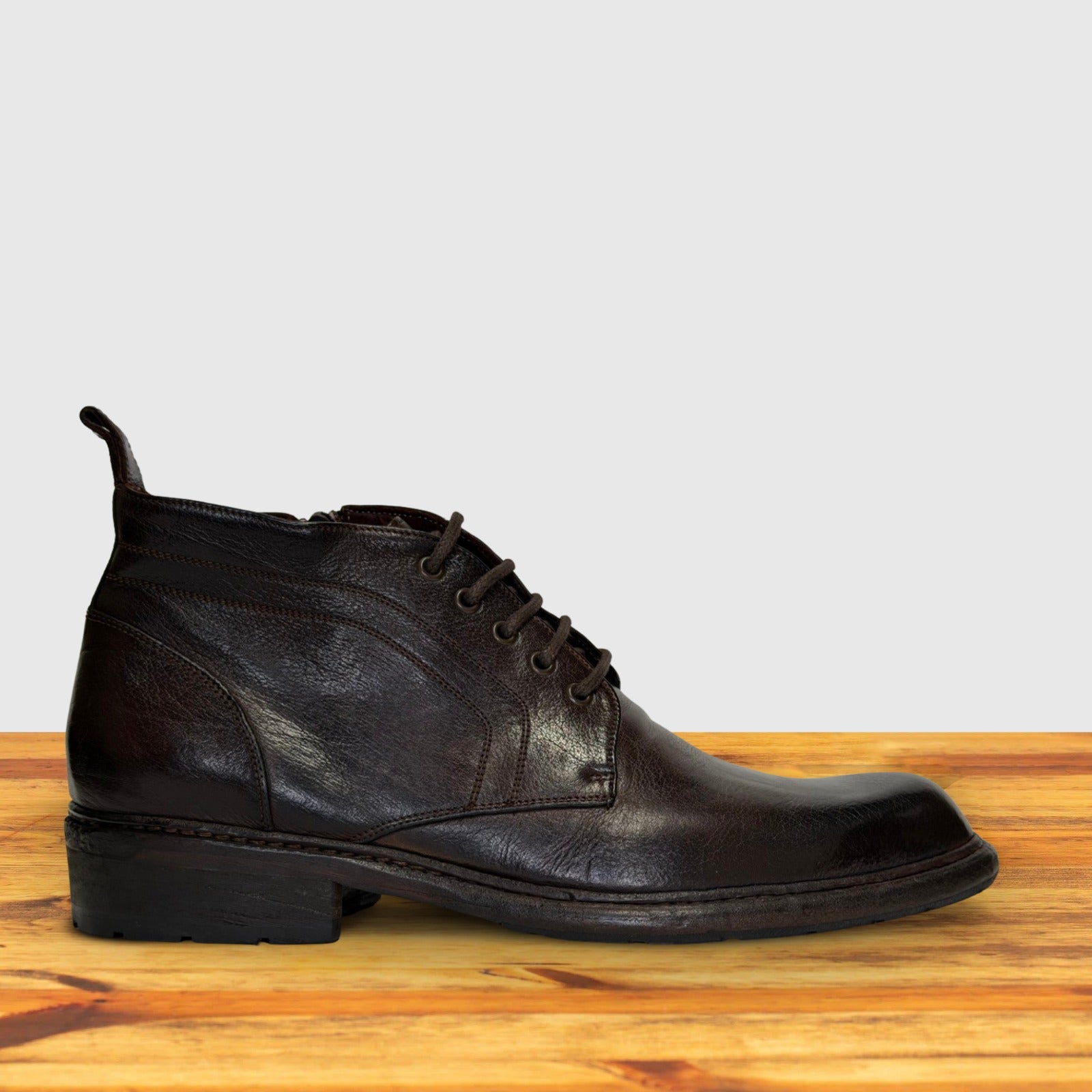 Dark Brown leather boot on a wooden surface with a light gray background