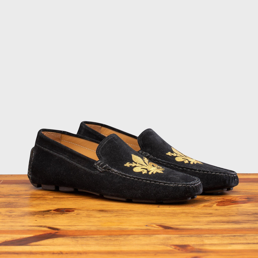 Pair of 5303 Calzoleria Toscana Black Venetian Suede Driver with gold embroidered fleur di lis on top of a wooden table