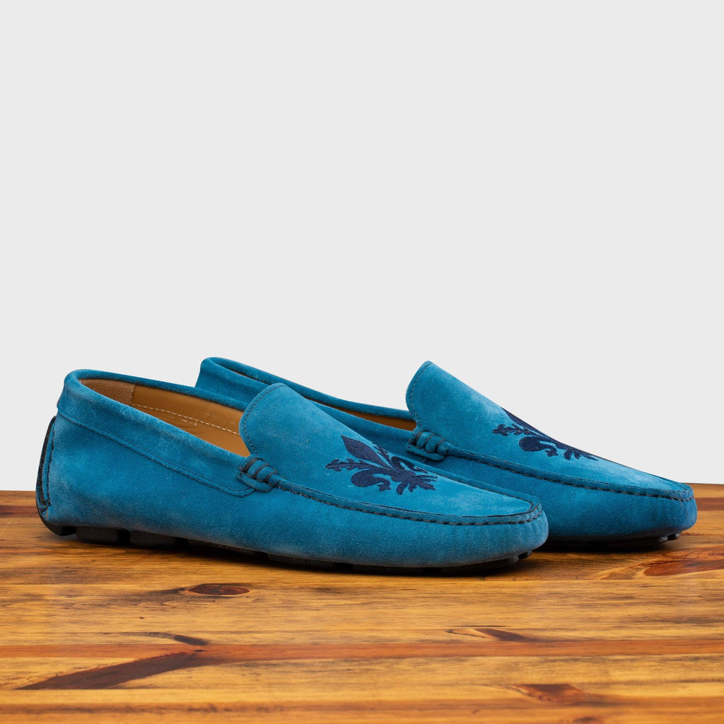Pair of 5303 Calzoleria Toscana Denim Blue Venetian Suede Driver with blue embroidered fleur di Lis on top of a wooden table