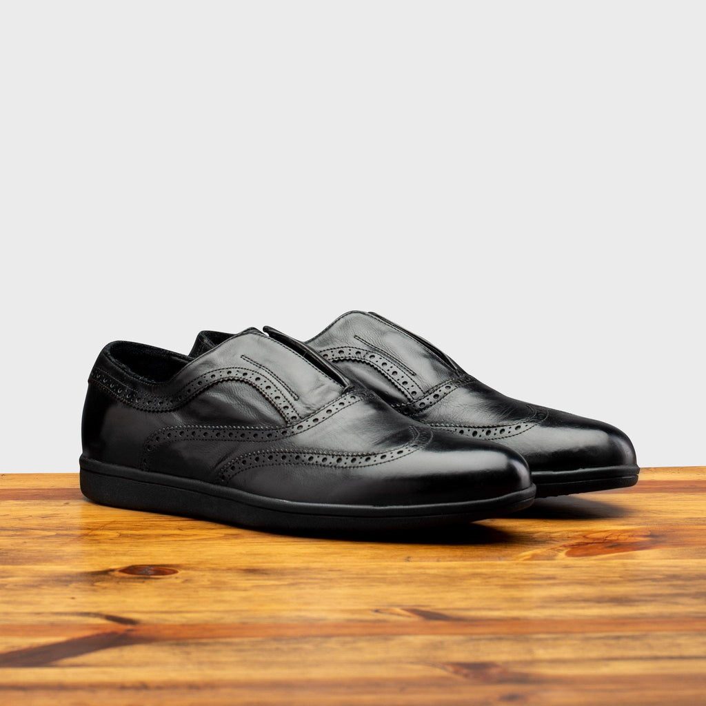 Pair of 7839 Calzoleria Toscana Black Elba Slip-On showing perforated wingtip details on top of a wooden table 