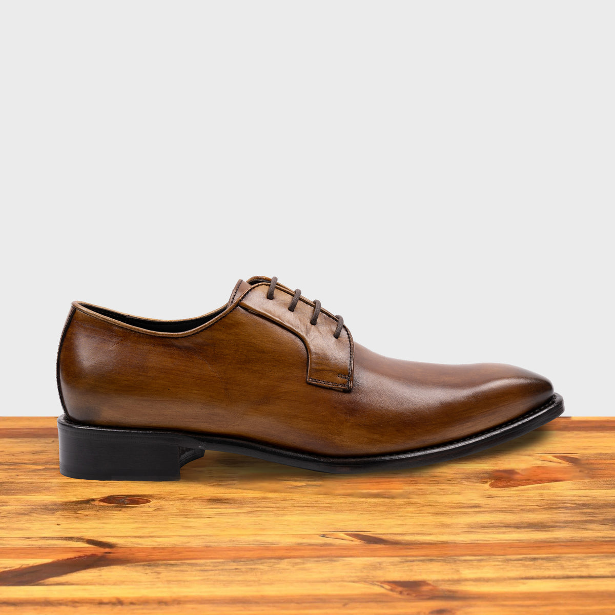 A Chestnut, wholecut blucher dress shoe with a lace-up closure, displayed on a wooden surface.