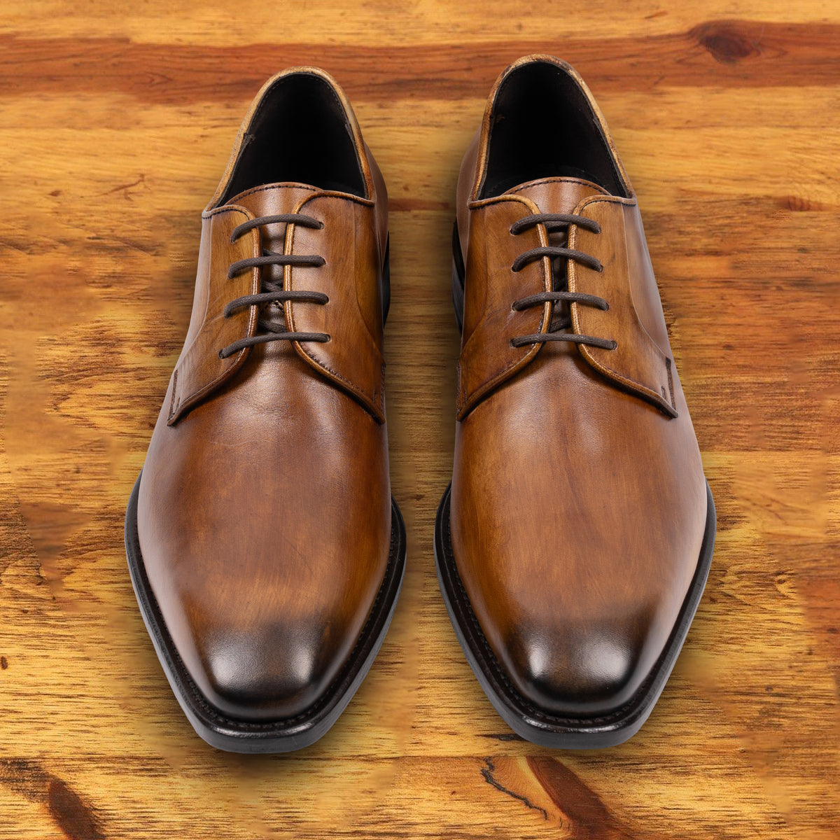 A Pair of Chestnut, wholecut blucher dress shoe with a lace-up closure, displayed on a wooden surface.