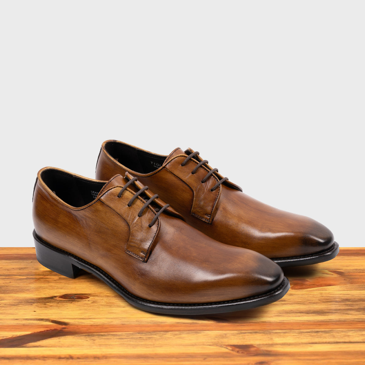 A Pair of Chestnut, wholecut blucher dress shoe with a lace-up closure, displayed on a wooden surface.